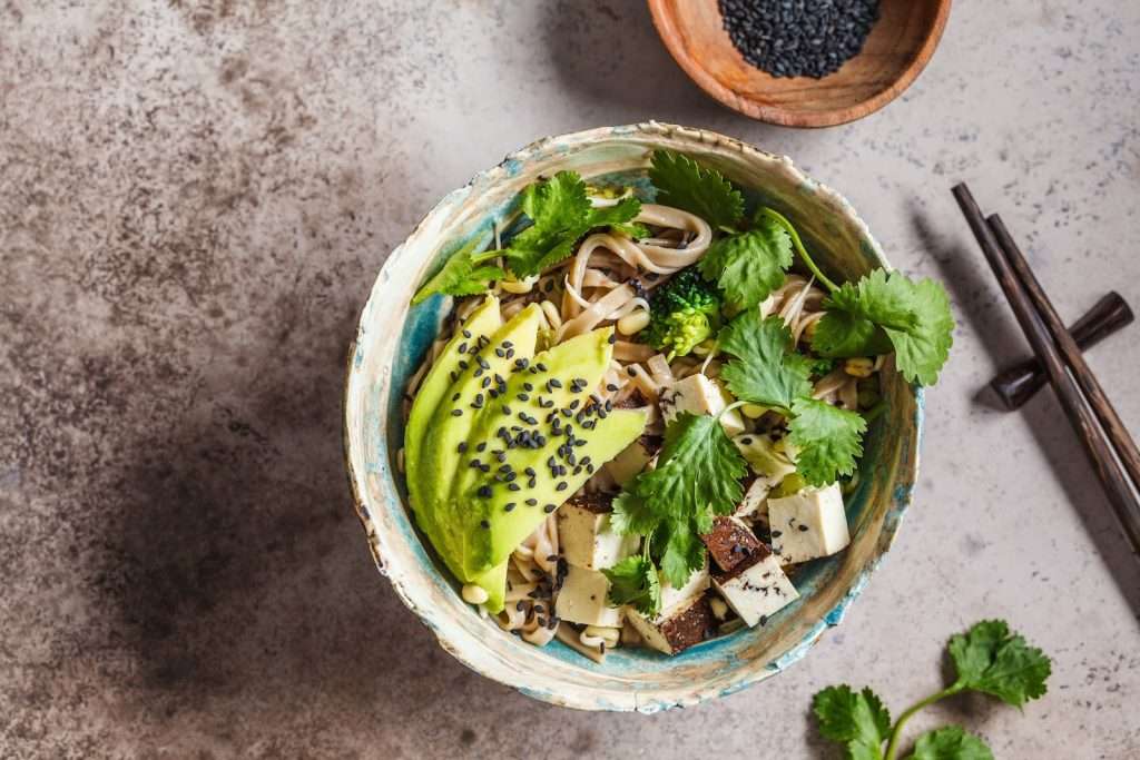 Vegan tofu noodle bowl with avocado, broccoli, cilantro, black sesame seeds, and chopsticks

