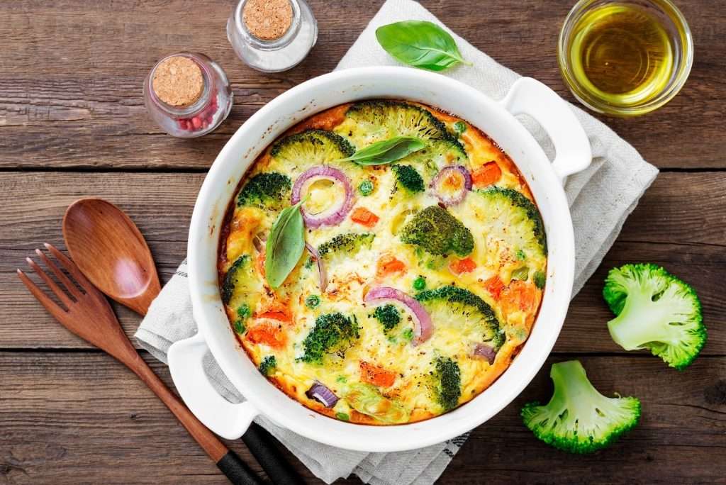 An easy vegan breakfast frittata baked in a white ceramic dish, loaded with fresh broccoli florets, red onions, carrots, and peas, served on a rustic wooden table.