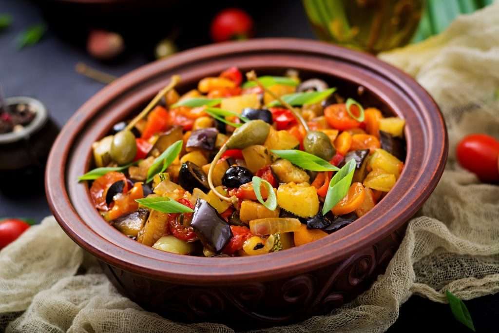 Vegan Mediterranean vegetable stew with eggplant, zucchini, tomatoes, olives, capers, and green onions in a clay bowl