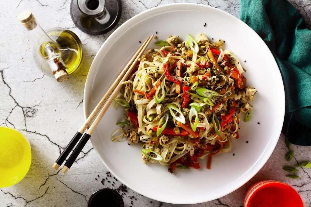 Vegan noodle stir-fry with tofu, red bell peppers, carrots, green onions, sesame seeds, and chopsticks in a white bowl