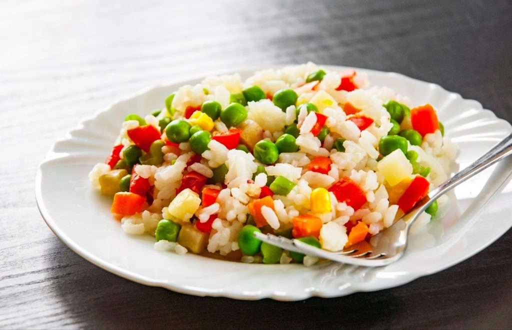 A plate of quick and easy vegan fried rice or seasoned vegetable rice pilaf mixed with green peas, diced carrots, corn, and red bell peppers, served on a white plate with a fork.