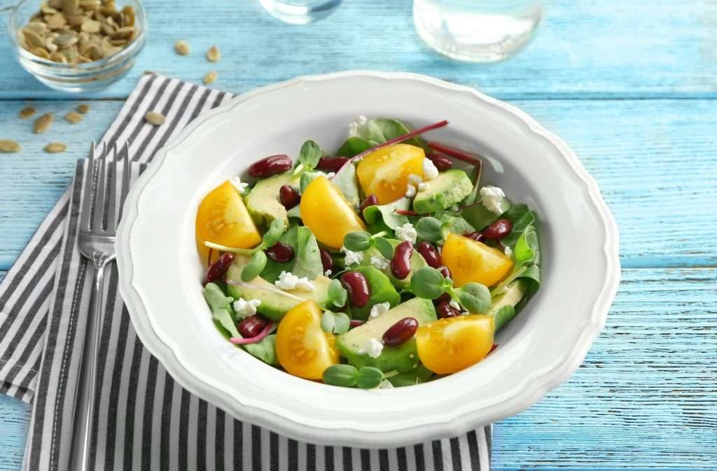 A quick and easy vegan avocado salad or healthy lunch bowl with yellow tomatoes, kidney beans, microgreens, and crumbled plant-based feta cheese, served in a white bowl on a blue wooden table.