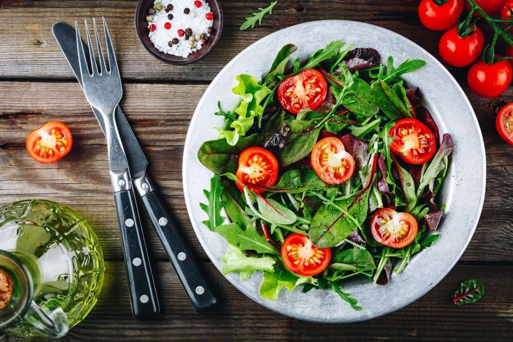 Fresh vegan green salad with cherry tomatoes, mixed leafy greens, arugula, olive oil, and seasoning on a rustic wooden table
